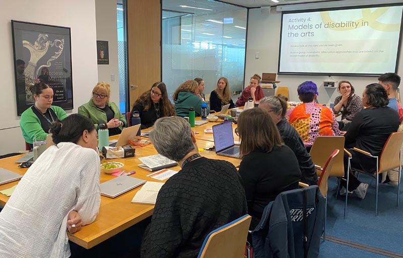 A group of Te Papa staff sit around a table, looking towards a screen that reads, "Models of disability in the arts"